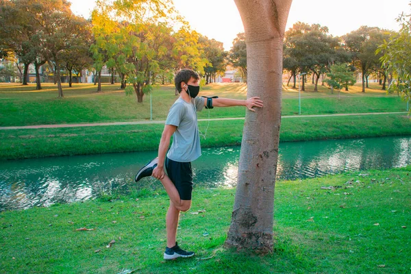 Spor kıyafetleri ve maske takmaya başlamadan önce yeşil bir parkta esneme hareketleri yapan genç bir adam..
