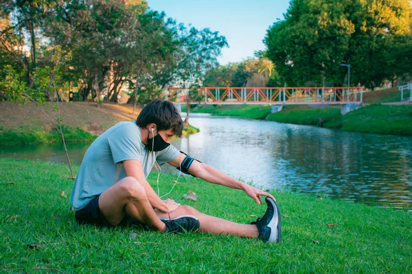 Spor kıyafetleri ve maske takmaya başlamadan önce yeşil bir parkta esneme hareketleri yapan genç bir adam..