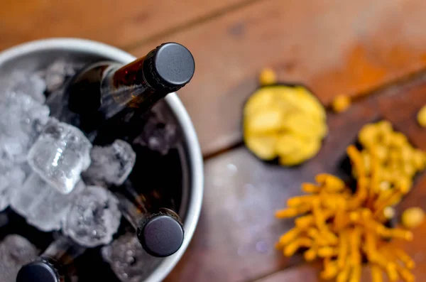 Black ice bucket with two beer bottles inside on a wooden table and with some savory snacks.