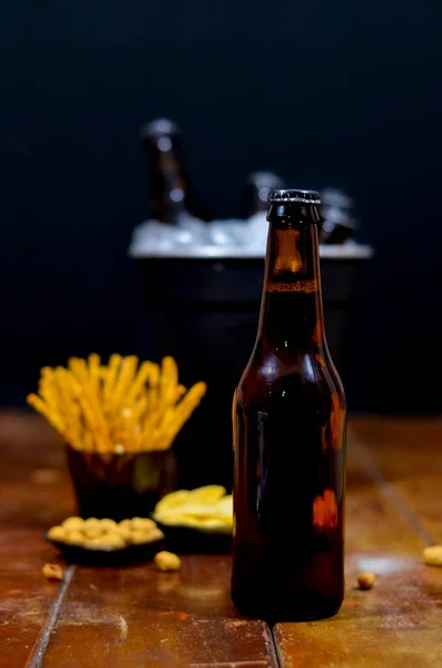 Ön planda ve arka planda bir şişe bira içinde bira şişeleri ve tuzlu atıştırmalıklar olan siyah bir buz kovası ahşap masa ve siyah arka planda..