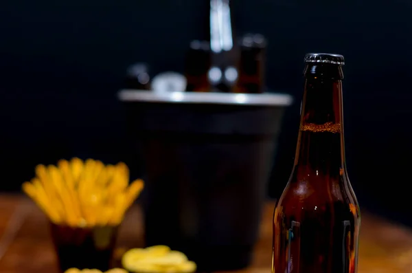 Ön planda ve arka planda bir şişe bira içinde bira şişeleri ve tuzlu atıştırmalıklar olan siyah bir buz kovası ahşap masa ve siyah arka planda..