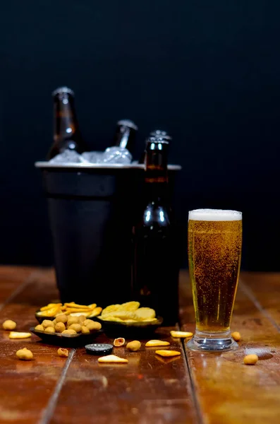 Ön planda bira dolu bir bardak, yanında bir şişe bira ve içinde bira olan siyah bir kova, ahşap bir masada lezzetli atıştırmalıklar..