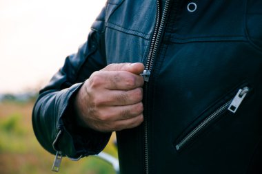 Yüzü görünmeyen, kot pantolon ve siyah ceket giyen, arkasında motosikletle ceketinin fermuarını çeken bir adam..