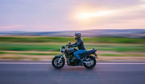 Siyah motosikletli bir adam, kot pantolon ve siyah ceket giymiş, arka planı bulanık bir şekilde yolda yürüyor..