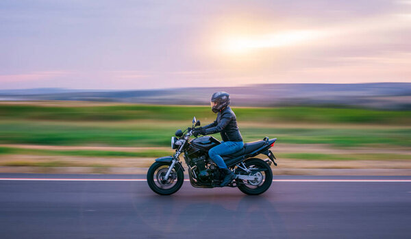 Man sitting on a black motorcycle, dressed in jeans and black jacket, moving on the road with the background out of focus.