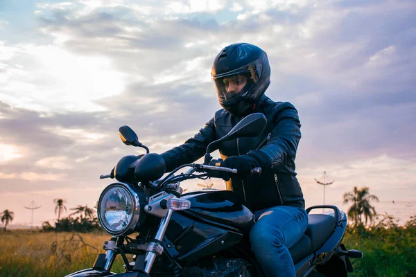 Siyah motosikletli bir adam, kot pantolon, siyah ceket ve bulutlu gökyüzü arka planı olan siyah bir kask giyiyor..