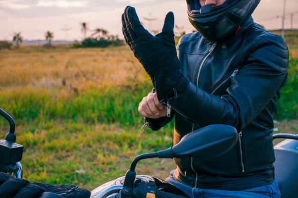 男人坐在一辆黑色摩托车上 穿着牛仔裤和黑色夹克 戴着黑色手套 背景是风景 — 图库照片