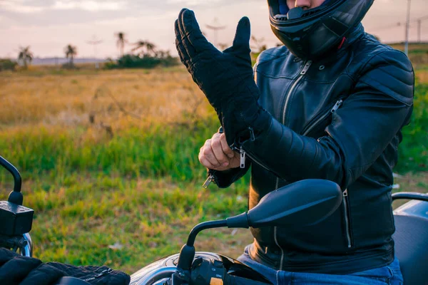 Siyah bir motosiklette oturan, kot pantolon ve siyah bir ceket giyen arka planda manzarası olan siyah eldivenler giyen bir adam..