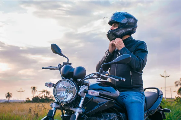 Motosiklette oturan, kot pantolon ve siyah ceket giyen, kaskını arka planda manzarayla bağlayan bir adam..
