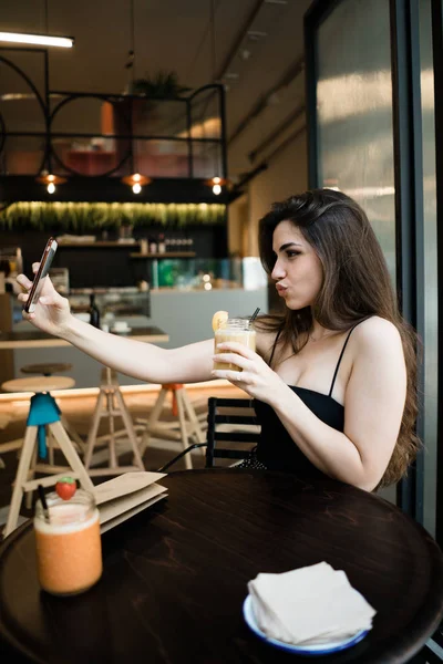 Şık genç esmer kadın oturma, koyu ahşap masa içecek ve alma selfie smartphone ön kamera Cafe arka plan bulanık dudaklarından somurtarak üzerinde tutarak yan görünüm 
