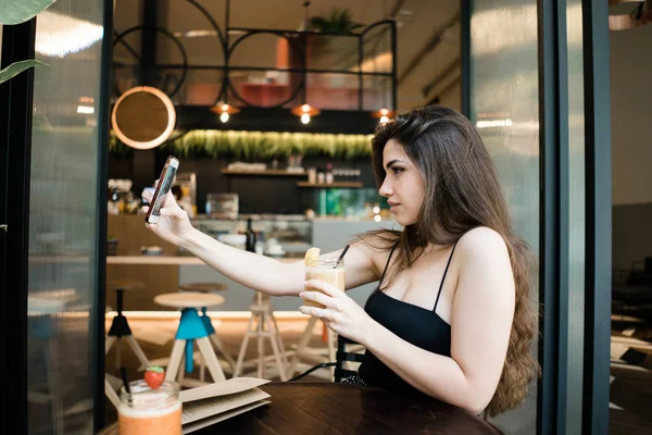 Smartphone ön kamera Cafe bulanık arka plan üzerinde üzerinde muhteşem genç esmer kadın oturma masada içecek kokteyl kavanoz ve alma resim ile yan görünüm