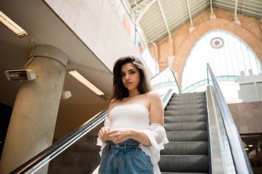 Zarif yürüyen merdiven üzerinde duran ve kameraya bakarak çıplak omuz ile beyaz yaz üst muhteşem genç esmer kadın 