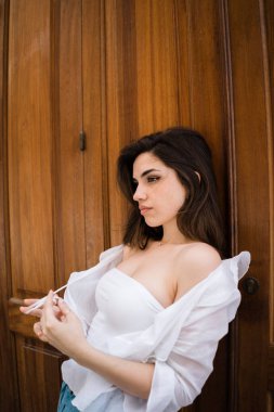 Güzel esmer kadın ahşap duvara yaslanmış ve uzak kayışlar, hafif bir ceket dokunmadan bakarak beyaz off--omuz üst yan görünüm 
