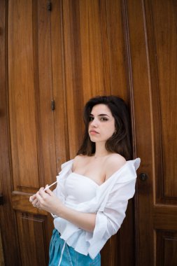 Çekici genç esmer Bayan ahşap kapıya yaslanmış ve beyaz off--omuz bluz askıları dokunmadan kameraya bakarak yaz giysileri 