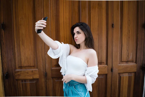 Gorgeous stylish female in summer off-the-shoulder clothes standing at wooden door and taking selfie on smartphone pouting lips