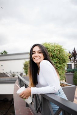 Güzel genç kadın yan görünüm gülümseyen ve kameraya teras korkuluk bulutlu gün üzerine eğilerek süre seyir taze içecek fincan ile