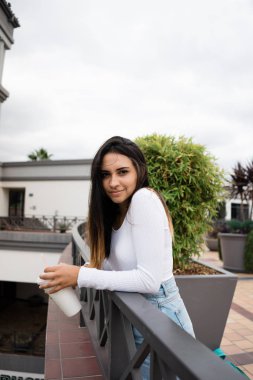 Oldukça genç bayan şık kıyafet sırıtışları ve bulutlu gün teras korkuluk üzerine eğilerek süre lezzetli içecek fincan holding yan görünüm