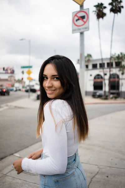 Yan görünüm şık genç ve güzel kadın kıyafeti neşeyle gülümseyen ve modern şehir Caddesi'nde kaldırımda duran kamera bakıyor