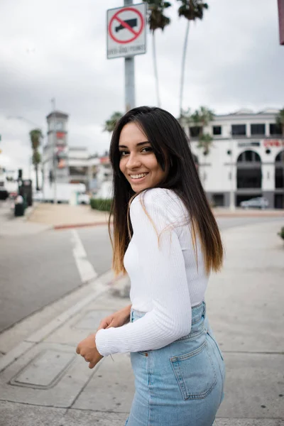 Çekici genç hanım gülümseyerek ve kamera güzel şehir sokak kaldırım üzerinde dururken bakarak şık casual kıyafet yan görünüm