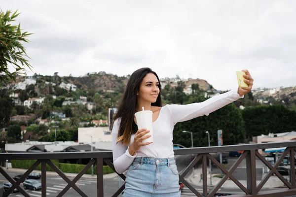 Gülümseyen ve teras korkuluk hills arka plan üzerinde ve bulutlu gün şehre yakın dururken selfie için poz taze içki Kupası ile büyüleyici genç erkek