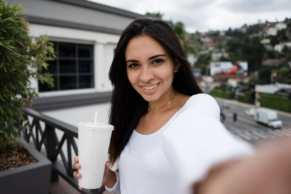 Sevimli genç kadın gülümseyerek ve şehir sokak arka plan üzerinde bina terasta selfie çekerken fotoğraf makinesi vasıl seyir taze içecek fincan ile