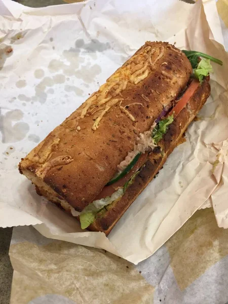 A toasted tuna fish grinder on herb and cheese bread unwrapped and sitting on the packaging