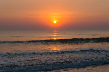 Parlak turuncu gökyüzü ve yumuşak deniz dalgaları üzerinde güneş ile plaj günbatımı manzara