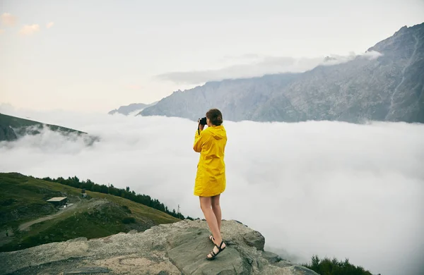 Una joven con un impermeable amarillo fotografía las montañas. Georgia. Verano. Agosto. Chica haciendo una sesión de fotos de la montaña . — Foto de Stock