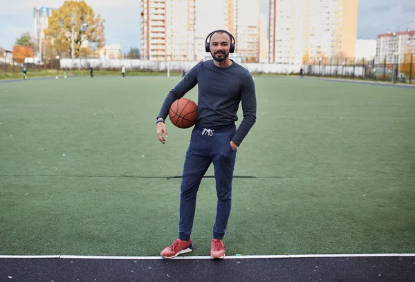 Atleta com uma bola de basquete no estádio. Jovem com fones de ouvido segurando uma bola de basquete — Fotografia de Stock