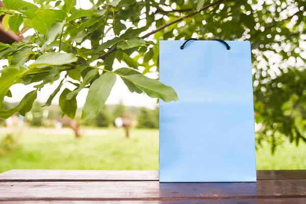 Blanco papieren zak staat op een houten tafel tegen de achtergrond van groene bladeren. Winkelconcept. Kopieerruimte. — Stockfoto