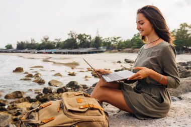 Genç kadın blogcu uzaktan bilgisayarla çalışıyor, interneti kullanıyor, deniz kıyısında, sahilde oturuyor. Seyahat ederken tatilde çalış. Monitöre bakıyorum. Serbest meslek kavramı. Yer kopyala