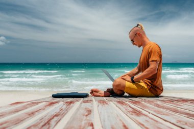 Güneşli bir günde deniz kenarında uzun saçlı genç bir adam laptopunu kullanarak çalışıyor, uzaktan bir işle uğraşıyor. Sarışın, serbest çalışan, internet üzerinden çalışan, iş seyahati yapan. Yaz tatili. Okyanusun kıyısındaki kumsalda ahşap bir patika. Yer kopyala