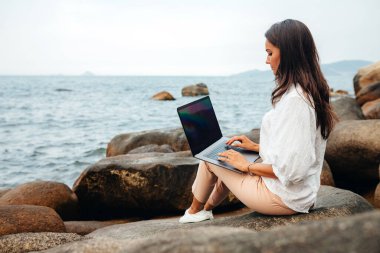 Genç bir kız dizüstü bilgisayar kullanarak çalışıyor, kayalık bir deniz kıyısında oturuyor. Seyahat ederken, blogcu, serbest çalışan, içerik planı hazırlayan, çevrimiçi çalışan bir iş kadını. Boşluğu kopyala
