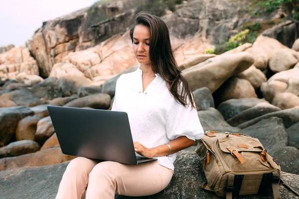 Young successful girl working using a laptop, sitting on a rocky seashore. Business woman working on remote work while traveling, blogger, freelancer, compiles content plan, work online. copy space