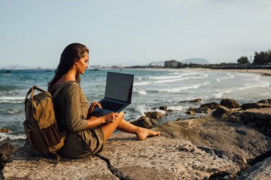 Deniz kenarında seyahat ederken dizüstü bilgisayarda çalışan genç ve güzel bir bayan serbest yazar, uzaktan çalışma konsepti. Deniz kenarında çalış. Modern teknolojiyi, interneti iletişim için kullanmak. Kayalık bir sahilde oturmuş, gülümseyerek çalışan bir kız.