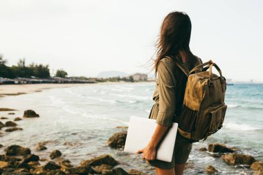 Kayalık bir deniz kıyısındaki bir kızın arkasından bir manzara, bir gezgin. Sırt çantalı ve elinde dizüstü bilgisayarla serbest çalışan bir işçi. Deniz manzaralı. Seyahat, tatil kavramı. Metin için yer, kopyala
