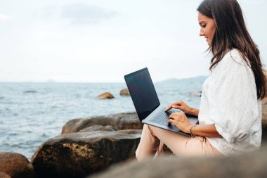 Uzak çalışma genç başarılı, gülümseyen kız dizüstü bilgisayar kullanıyor, kayalık bir deniz kıyısında oturuyor. Seyahat ederken çalışan iş kadını, blogcu, serbest çalışan, içerik planı hazırlayan, çevrimiçi çalışan. Boşluğu kopyala