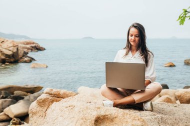 Blogcu kız dizüstü bilgisayar kullanıyor, kayalık bir deniz kıyısında oturuyor. Seyahat ederken uzaktan çalışan iş kadını, serbest çalışan, içerik planı hazırlayan, çevrimiçi çalışan. Boşluğu kopyala