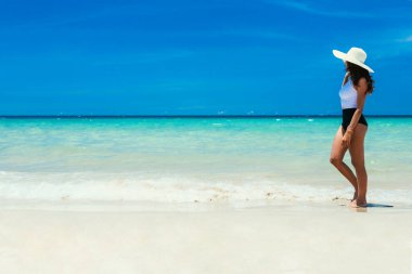 Deniz, sahil, kumsal, kumlu bir sahil boyunca yürüyen güzel bir kız. Denizde berrak berelenmiş bir bereli su, hasır bir şapka içinde. Sakin deniz ve arkada açık mavi gökyüzü. Dinlenmek, seyahat etmek. Yer kopyala