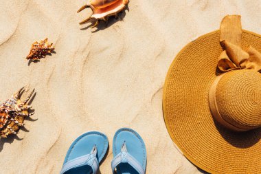Üzerinde deniz kabukları olan hasır bir şapka, kumlu bir kumsalın kıyısında, şapkanın yanında terlikler. Dinlenme ve denizde seyahat kavramı. Yer kopyala