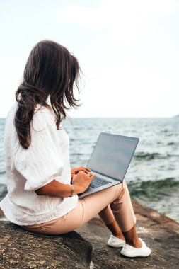 Genç başarılı kız dizüstü bilgisayar kullanarak çalışıyor, kayalık bir deniz kıyısında oturuyor. Uzaktan kumandalı iş kadını, blogcu, serbest çalışan, içerik planı hazırlayan, çevrimiçi çalışan. Boşluğu kopyala