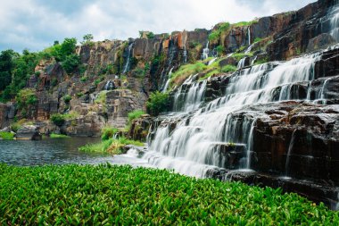 Popüler bir turistik yerde bir şelale. Kayalık çıkıntılar boyunca uzanan güçlü bir su akıntısı yeşillik ile kaplanmış bir göle dökülüyor. Uzun pozlama çekimi
