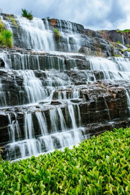 Popüler bir turistik yerde bir şelale. Kayalık çıkıntılar boyunca uzanan güçlü bir su akıntısı yeşillik ile kaplanmış bir göle dökülüyor. Uzun pozlama çekimi