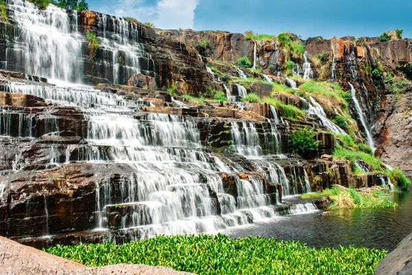 Popüler bir turistik yerde bir şelale. Kayalık çıkıntılar boyunca uzanan güçlü bir su akıntısı yeşillik ile kaplanmış bir göle dökülüyor. Uzun pozlama çekimi
