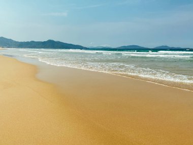Tropik tatil konsepti. Uzun ve boş bir deniz sahilinin tropikal sahilinde yumuşak uzun dalgalar. Sakin dalgalı Sandy Oceanic plajı. Arka planda beyaz bulutlar olan güzel mavi gökyüzü. Karayip kıyısında deniz köpüğü