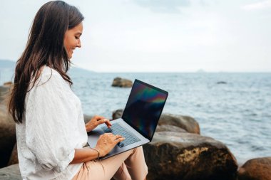 Serbest meslek kavramı. Kucağında dizüstü bilgisayarla gülümseyen serbest çalışan bir kız deniz kenarındaki bir kayanın üzerinde otururken ofis dışında çalışıyor. Temiz havada uzaktan çalışma, modern işçi. Boşluğu kopyala