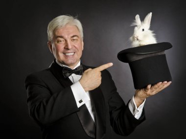 Yaşlı bir gri saçlı prestidigitator siyah smokin siyah bir şapka ve bir tavşan ile magic tricks karanlık bir arka plan üzerinde gösterir.