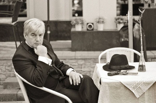 A grey-haired elderly mobster sitting in a summer cafe. Agrey-haired elderly mobster in a black suit with a gun sitting in a summer cafe against a vintage background.