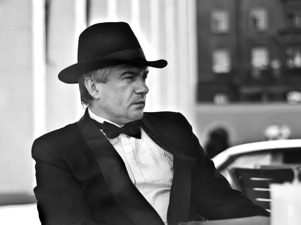 A grey-haired elderly mobster sitting in a summer cafe. A grey-haired elderly mobster in a black suit and a black hat sitting in a summer cafe against a black-and-white background.