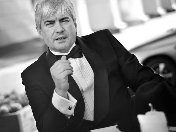 A grey-haired elderly mobster sitting in a summer cafe. A grey-haired elderly mobster in a black suit sitting in a summer cafe against a black-and-white background.